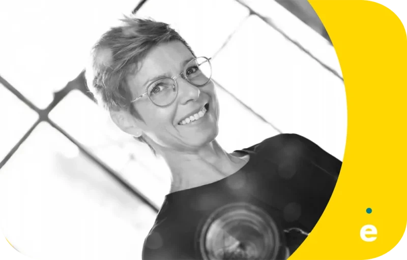 Une personne souriante portant des lunettes rondes est photographiée en noir et blanc devant une fenêtre avec un design graphique jaune.