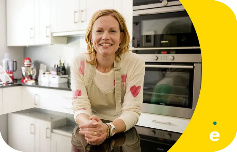 Une personne souriante en tablier se tient dans une cuisine moderne avec des appareils électroménagers en arrière-plan.