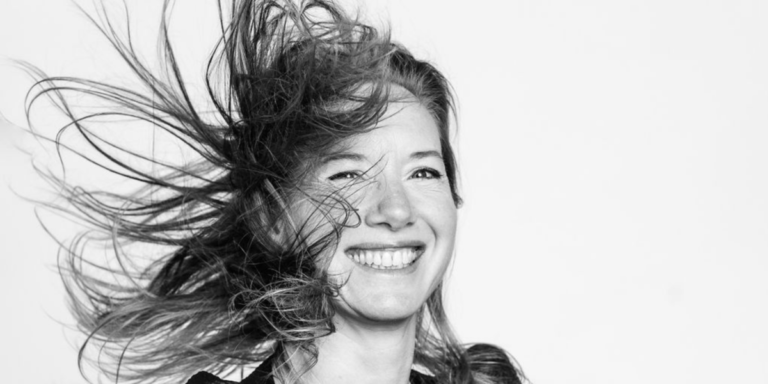 Une personne souriante avec les cheveux ébouriffés, en noir et blanc, sur fond uni.