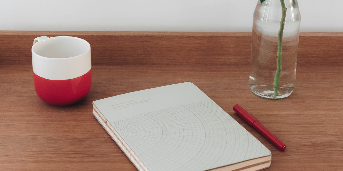 Tasse rouge et blanche, cahier à motifs géométriques, stylo rouge et vase avec une rose sur une table en bois.