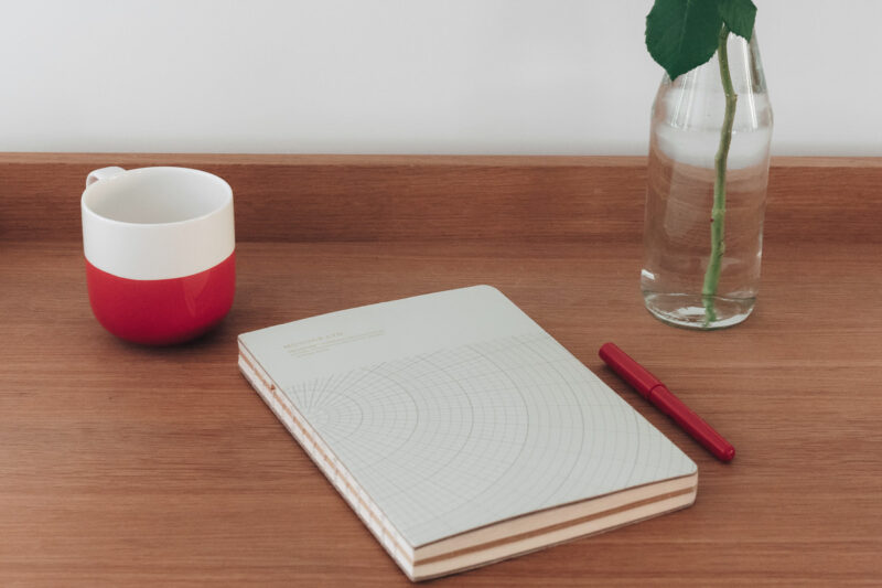 Tasse rouge et blanche, cahier à motifs géométriques, stylo rouge et vase avec une rose sur une table en bois.