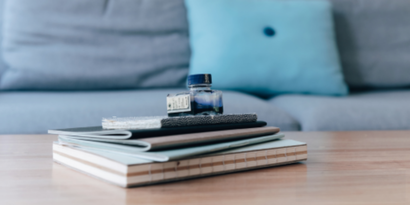 3 étapes pour faire décoller ton entreprise Des carnets empilés sur une table en bois avec un flacon d'encre bleu, en arrière-plan un canapé gris avec un coussin bleu.