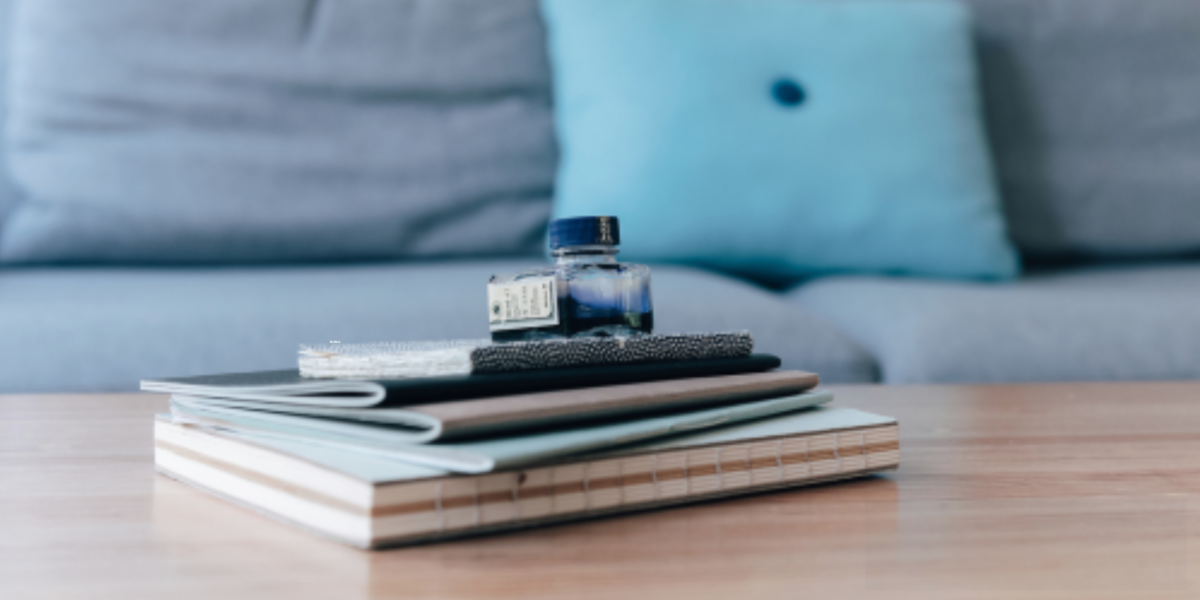 Des carnets empilés sur une table en bois avec un flacon d'encre bleu, en arrière-plan un canapé gris avec un coussin bleu.