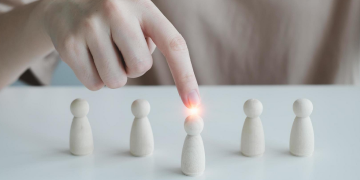 Une main pointe une figurine en bois parmi cinq alignées sur une table blanche, avec une lumière brillante sur celle-ci.