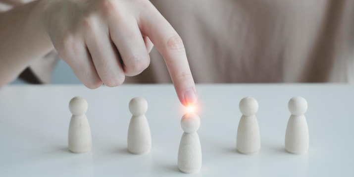 Une main pointe une figurine en bois parmi cinq alignées sur une table blanche, avec une lumière brillante sur celle-ci.