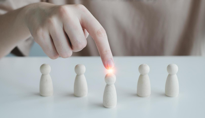 crédit : vecteezy.com Une main pointe une figurine en bois parmi cinq alignées sur une table blanche, avec une lumière brillante sur celle-ci.