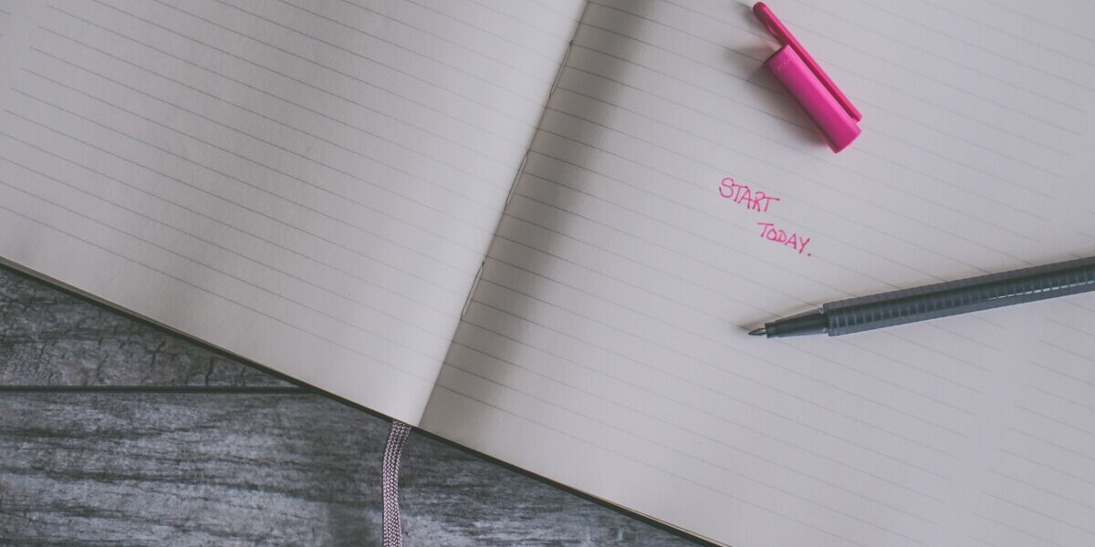 Carnet ouvert avec lignes, stylo noir et capuchon rose sur une table en bois grise, texte "START TODAY" écrit en rose.