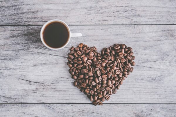 Une tasse de café noir à gauche et des grains de café formant un cœur à droite sur une table en bois gris.