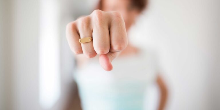 Une personne tend le poing vers l'objectif, portant une bague dorée sur fond flou.