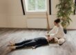 Une personne allongée sur un tapis de yoga reçoit un soin énergétique d'une autre personne dans une pièce lumineuse avec une plante verte.