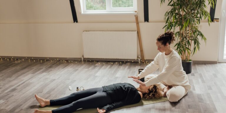 Une personne allongée sur un tapis de yoga reçoit un soin énergétique d'une autre personne dans une pièce lumineuse avec une plante verte.