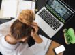 Une personne assise à un bureau avec un ordinateur portable, un cahier ouvert, un téléphone et des lunettes vertes posées à côté.