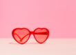 Lunettes de soleil rouges en forme de cœur posées sur une surface beige avec un fond rose uni.