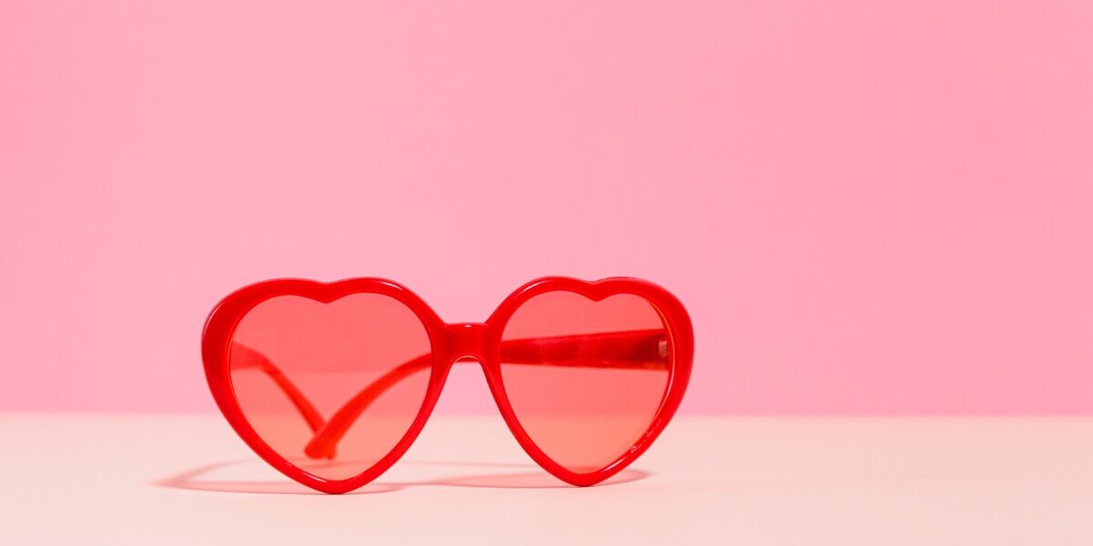 Lunettes de soleil rouges en forme de cœur posées sur une surface beige avec un fond rose uni.