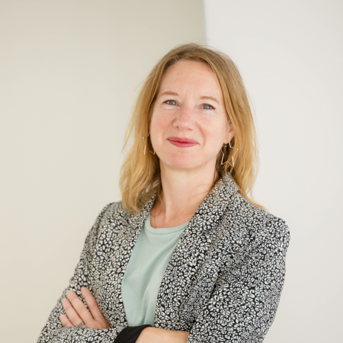 Une femme aux cheveux blonds porte un blazer à motifs noirs et blancs, avec un fond beige et blanc uni.