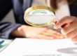 Une personne en costume examine un document avec une loupe dorée, posée sur une table avec des stylos colorés.