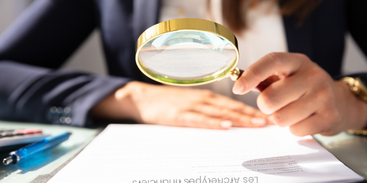 Une personne en costume examine un document avec une loupe dorée, posée sur une table avec des stylos colorés.