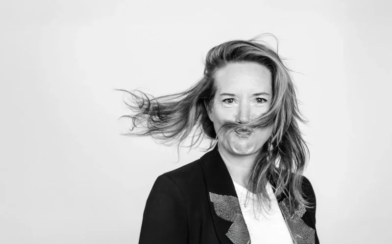 Une personne aux cheveux longs souffle sur ses mèches, vêtue d'une veste noire, sur un fond blanc uni.