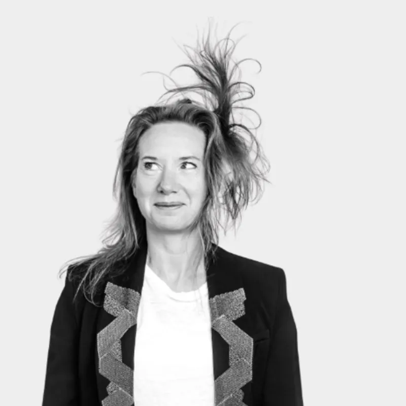 Une femme en noir et blanc avec les cheveux ébouriffés porte une veste noire et un t-shirt blanc sur fond gris clair.