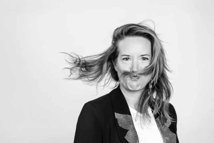portrait d'une femme : Delphine Terrasson aux cheveux longs souffle sur ses mèches, vêtue d'une veste noire, sur un fond blanc uni.