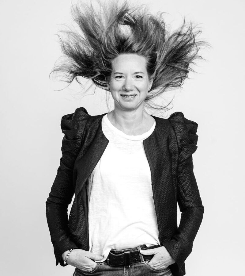 une femme face caméra heureuse les cheveux en l'air vétue d'une veste noire et tee-shirt blanc, regardant face caméra